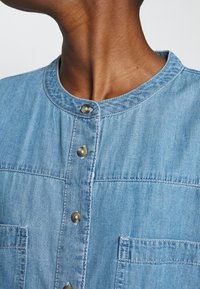 Hellblaue Jeanshemd mit runder Ausschnitt, zwei Brusttaschen und Knopfverschluss. Der Stoff hat eine weiche Textur und subtile Nähdetails.