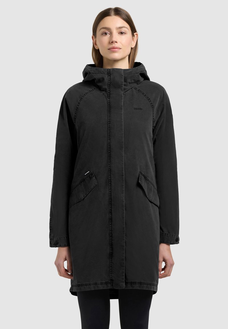 Femme portant un long manteau noir à capuche avec des poches à rabat à l'avant, debout devant un fond gris clair uni.