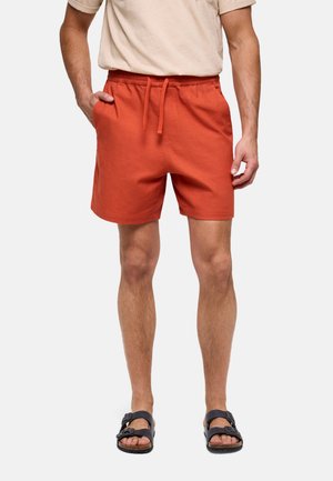 Hombre lleva pantalones cortos de cuerda color naranja quemado, camiseta beige y sandalias negras con hebilla, de pie con una mano en el bolsillo.