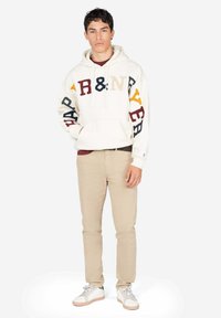 Sudadera crema con letras multicolores en las mangas, bolsillo frontal y cordón. Combinada con pantalones beige y zapatillas blancas.
