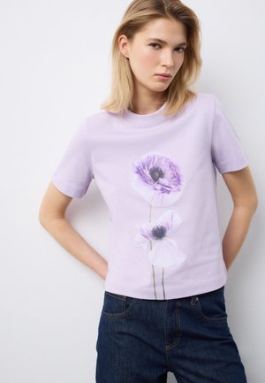 Giovane donna con indosso una maglietta lilla chiaro con la stampa di due grandi fiori viola, abbinata a jeans blu scuro, in piedi contro una parete bianca liscia.