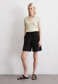 Beige Poloshirt mit kurzen Ärmeln und V-Ausschnitt, kombiniert mit lockeren schwarzen Shorts bis zum Knie und schwarzen Sandalen mit einer Schnallenakzent.