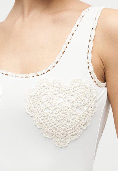 Κοντινή λήψη λευκού αμάνικου τοπ με πλεκτή διακόσμηση σε σχήμα καρδιάς και περιμετρική ραφή στο ντεκολτέ και την περιοχή της μασχάλης.