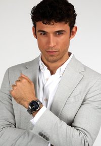 Silverfärgad rostfritt stål klocka med en svart urtavla, meshnätsband och detaljerade markörer, som bärs på ett handled. Modell i en ljusgrå kostym.