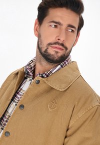 Veste en velours côtelé marron avec un grand col, présentant des boutons sombres et un logo d'ancre brodé. En dessous, une chemise à carreaux est visible.