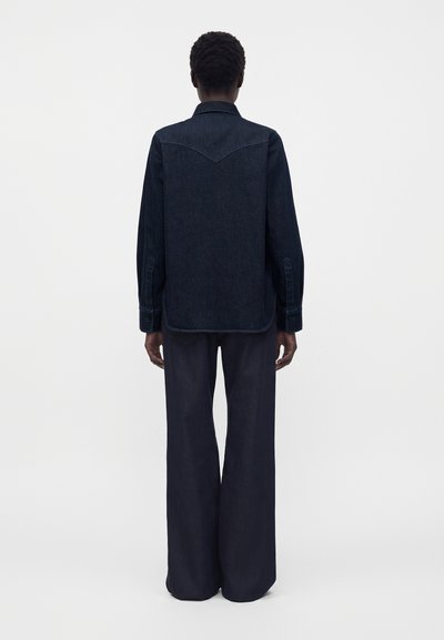Personne vue de dos portant une chemise en jean foncé à manches longues et un pantalon large ample foncé, debout devant un fond uni.