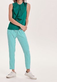 Teal mouwloos top met gerimpeld voorkantdetail, gecombineerd met teal en witte gestreepte broeken. Schoenen zijn witte sneakers met subtiele accenten.