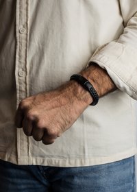 Brățară din piele împletită neagră cu un închizător metalic neted, purtată pe un încheietură. Fundalul prezintă o cămașă ușor colorată, cu nasturi.