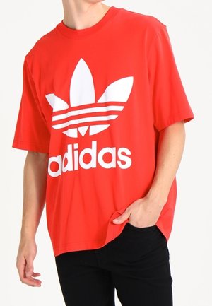 Osoba w czerwonym T-shircie Adidas z dużym białym logo trefoil i napisem, z ręką w kieszeni, na jednolitym tle.