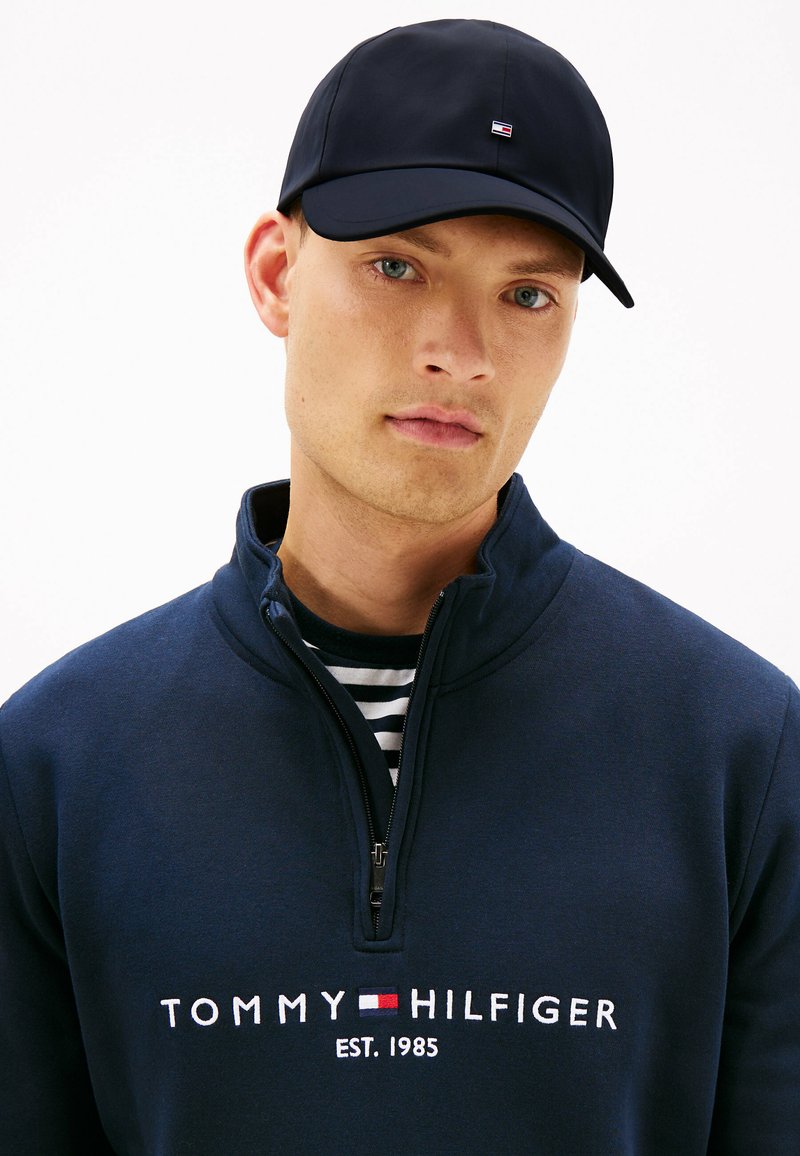 Gorra de color azul marino oscuro y sudadera con cremallera con el logo "TOMMY HILFIGER"; la sudadera tiene un cuello alto y una camisa de rayas por debajo.