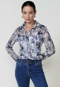 Una modella posa con sicurezza, indossando una camicia a maniche lunghe a fantasia floreale e jeans blu, con uno sfondo neutro e gioielli semplici.