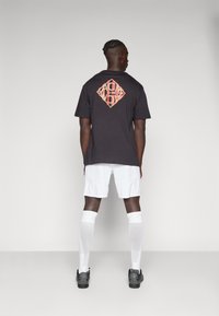 Svart bomull t-shirt med en färgglad diamantgrafik på ryggen, kombinerad med vita shorts och knästrumpor, bärs med svarta skor.