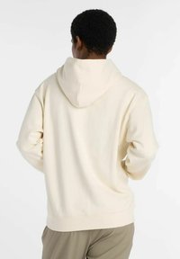 Persona con cabello corto y rizado, vistiendo una sudadera color crema y pantalones beige, de pie con las manos en las caderas y mirando hacia atrás.