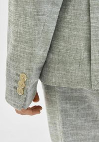 Ljusgrå linnéblazer ärm med fyra beige knappar, som visar en texturerad tyg och en skräddarsydd passform.