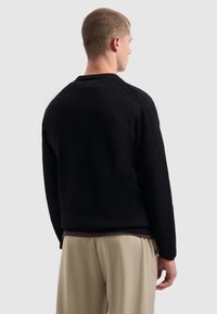 Man die zich omdraait en een zwart langemouwen shirt en beige broek draagt, tegen een effen lichtgrijze achtergrond.