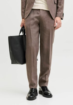 Persona vestida con pantalones y chaqueta marrones, sosteniendo un bolso tote de cuero negro, calzando zapatos de vestir de cuero negro.