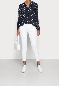 Camicia a pois blu navy, maniche lunghe, stile button-down, abbinata a jeans bianchi slim-fit e sneakers bianche, mentre tiene una piccola borsa bianca.