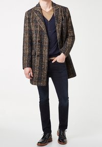 Kabát z vlněného materiálu s houndstooth vzorem v tmavě hnědé, béžové a námořnické modři, vrstvený přes námořnický svetr a dobře padnoucí tmavé džíny, doplněný o černé boty.