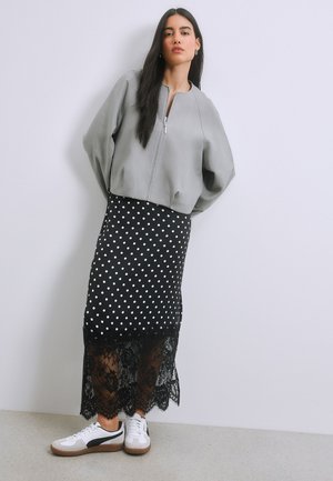 Nainen, jolla on pitkät tummat hiukset, harmaa vetoketjullinen takki, mustavalkoinen pilkkkuhame, jossa on pitsireunus, ja valkoiset lenkkarit valkoista seinää vasten.