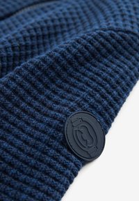 Tissu tricoté bleu foncé avec une texture gaufrée et un patch rond noir orné d'un logo ou emblème stylisé cousu dessus.