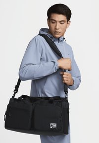 Nike treningsbag har et svart stoffutsiden, doble topphåndtak, justerbar skulderstropp, flere glidelåslommer og meshdetaljer.