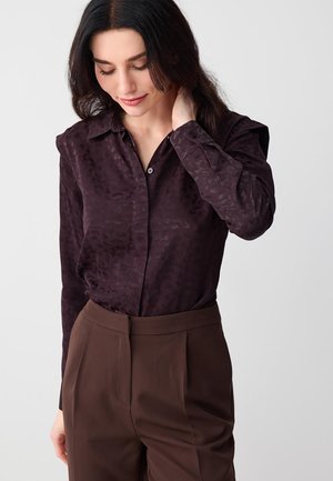 Femme aux cheveux foncés portant une chemise à manches longues à motifs foncés rentrée dans un pantalon marron taille haute, regardant vers le bas avec la main sur la nuque.