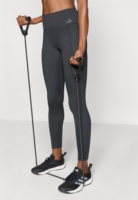 Σκούρο γκρι ριμπ κολάν με ψηλή μέση, σε συνδυασμό με μαύρα αθλητικά παπούτσια και λαστιχένιες ταινίες αντοχής, με μαύρες λεπτομέρειες.