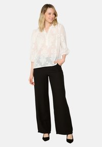 Blouse blanche transparente avec un décolleté en V, une patte de boutonnage et des manches volumineuses, assortie à un pantalon noir à jambes larges et des escarpins noirs.