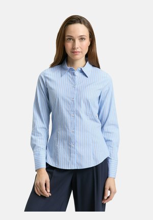 Mujer con una camisa azul claro ajustada de botones y finas rayas verticales rojas, y pantalones azul marino oscuro, posando frente a un fondo liso.