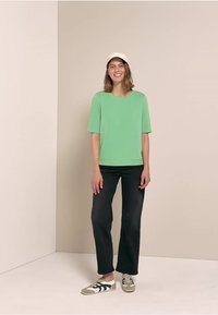 Chemise verte à manches courtes associée à un pantalon noir à jambes larges et des baskets grises avec des rayures vertes. Le modèle porte une casquette blanche.