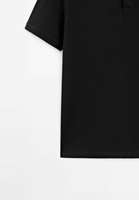 Polo shirt noir à manches courtes sur fond blanc, montrant la manche droite et la zone inférieure du torse avec une texture de tissu visible.