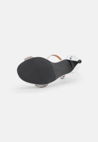 Sandały na przezroczystym obcasie z czarną podeszwą, przezroczystym wierzchem i ozdobnymi srebrnymi kwiatowymi akcentami. Posiadają cienki pasek wokół kostki dla wsparcia.