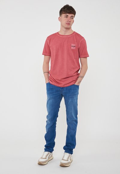 T-shirt rossa a maniche corte con una piccola tasca sul petto, abbinata a jeans blu e scarpe sportive di colore chiaro. Lo sfondo semplice migliora la visibilità.