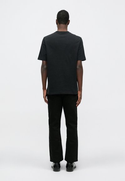 Tricou negru din bumbac cu guler rotund, asortat cu pantaloni negri drepti și pantofi negri, văzut din spate pe un fundal deschis.