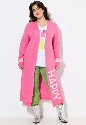 Maglione lungo rosa con apertura frontale, testo bianco "HAPPY" su un lato, polsini a righe a contrasto; indossato sopra una camicia bianca e pantaloni verdi. Stivali argentati.