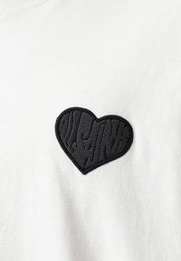 Camiseta de algodón blanca con un parche de corazón bordado en negro, con detalles texturizados y letras en forma de dientes de sierra dentro de la forma del corazón.