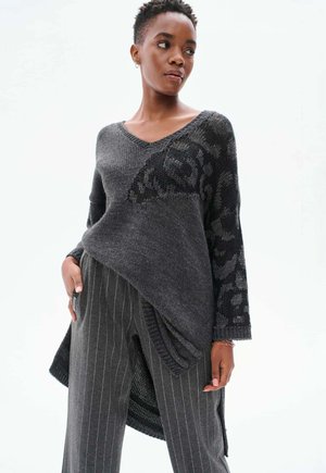 Asymmetrischer grauer Strickpullover mit schwarzen gemusterten Akzenten und einem V-Ausschnitt, kombiniert mit grauen gestreiften Hosen. Strukturierter Stoff mit lockerer Passform.