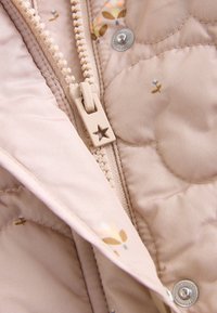 Tissu beige matelassé pour veste avec un curseur de fermeture éclair en forme d'étoile et petits motifs floraux, ainsi qu'un bouton pression en métal.