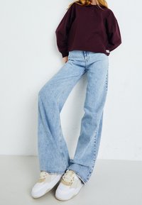 Tmavě červená polokošile s dlouhým rukávem, límečkem a knoflíky, ve světle modrých džínách s širokými nohavicemi a bílých teniskách s béžovými akcenty.