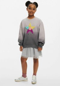 Grauer Sweatshirt mit Farbverlauf und einem mehrfarbigen Sterndesign, kombiniert mit einem mehrlagigen grauen Tüllrock und silbernen Sneakern.