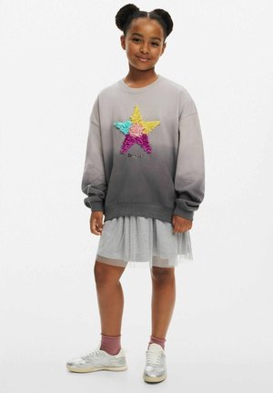 Grå gradiente sweatshirt med en mångfärgad stjärndesign, i kombination med en lager-på-lager grå tyllkjol och silverfärgade sneakers.