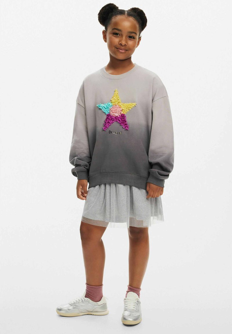 Grauer Sweatshirt mit Farbverlauf und einem mehrfarbigen Sterndesign, kombiniert mit einem mehrlagigen grauen Tüllrock und silbernen Sneakern.