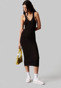 Vestito midi nero, aderente e a coste con scollatura a V profonda. Indossato con sneakers bianche e una borsa gialla con design strutturato e cuciture a contrasto.