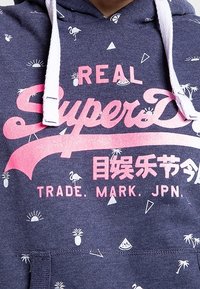 Felpa blu marina con testo grafico bianco e rosa "REAL SuperDry" e piccoli motivi tropicali, tra cui fenicotteri e ananas.