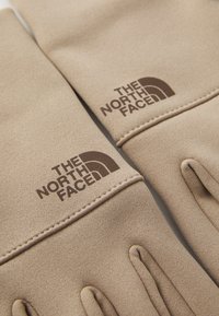 Beige hansker laget av et mykt, teksturert materiale med sydd accenter. Har en brun logo av "The North Face" på hvert håndledd.