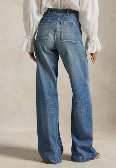 Personne portant un jean bleu taille haute à jambes larges et un chemisier blanc à manches longues avec des poignets froncés, debout sur un fond neutre.