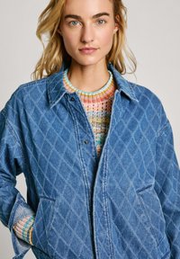 Giubbotto di jeans blu trapuntato con chiusura a scatto, dotato di colletto appuntito e tasche laterali. Indossato sopra un maglione lavorato a maglia multicolore.