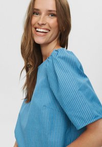 Modra prugasta bluza s kratkimi rokavi, ki ima teksturirano tkanino in majhno žep na sprednji strani. Preprost, sodoben dizajn.