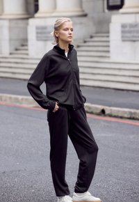 Schwarze Trainingsjacke mit Reißverschluss und Seitentaschen sowie passende Hose. Modell trägt weiße Turnschuhe. Glatte Textur, lockere Passform, minimalistisches Design.