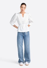 Camicia bianca con scollo a V e maniche a sbuffo, cinchata in vita con un nastro. Abbinata a jeans a gamba larga azzurri e sneaker bianche.